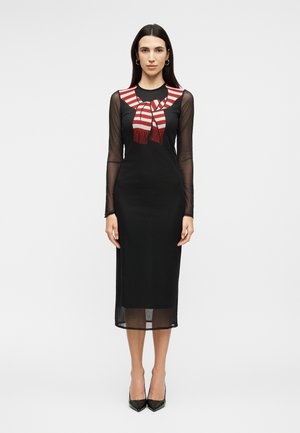 Nő fekete átlátszó hosszú ujjú midi ruhában, piros-fehér csíkos sálrészlettel a mellrészen, egyszerű háttér előtt állva.