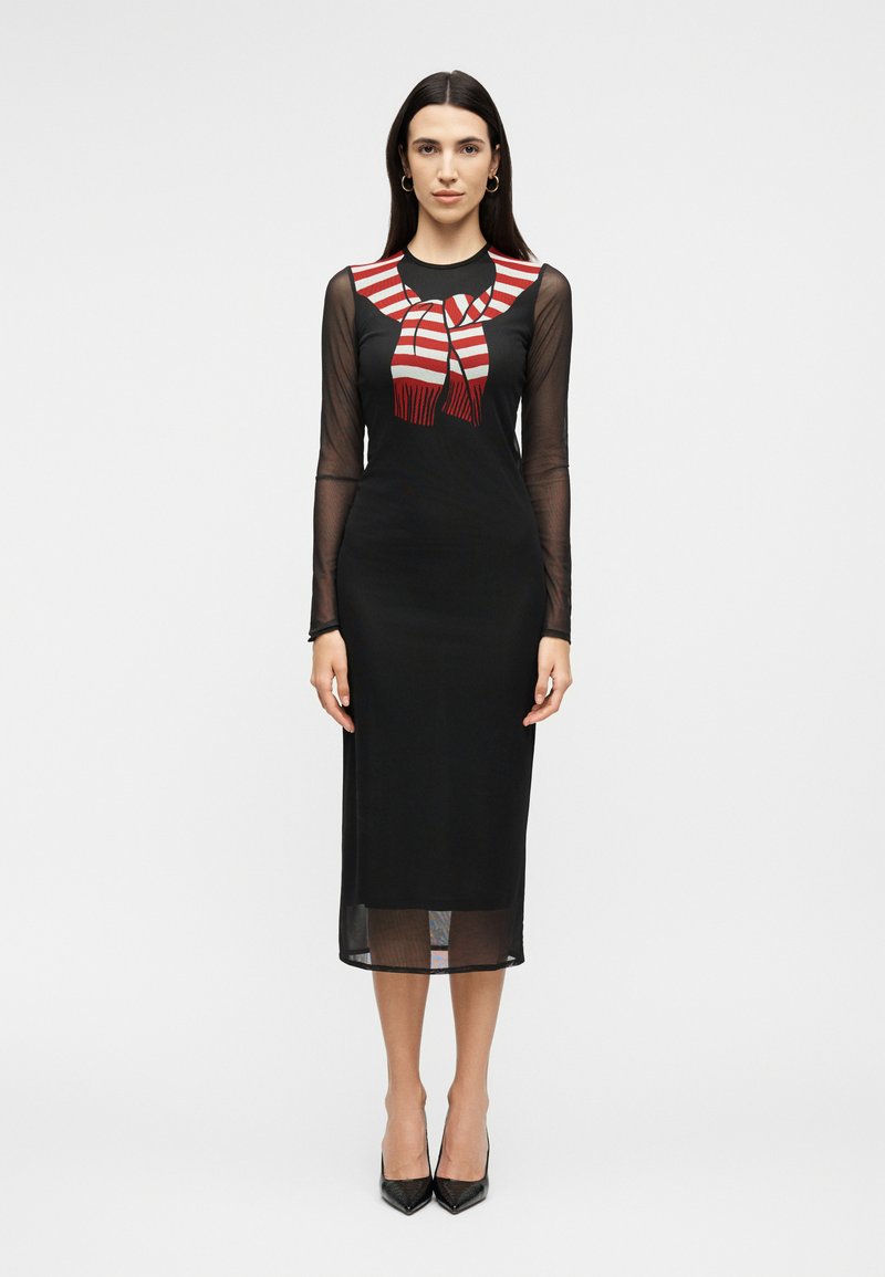 Kobieta w czarnej, prześwitującej, midi sukience z długim rękawem, z dodatkiem czerwono-białego szalikowego wzoru na wysokości klatki piersiowej, stojąca na jednolitym tle.