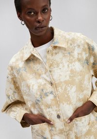 Lichte, beige denimjacket met blauw spetterpatroon, knoopsluiting en zijzakken. Stof heeft een licht gestructureerde afwerking.
