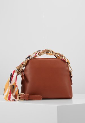 Bruine leren handtas met handvat van gevlochten multicolor sjaal en afneembare riem, rustend op een wit oppervlak tegen een grijze achtergrond.