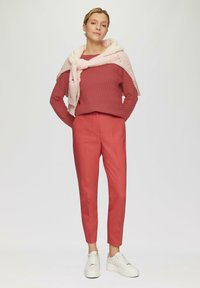 Koralfarbener gerippter Pullover, kombiniert mit einem rosa Strickschal, getragen zu maßgeschneiderten koralfarbenen Hosen und weißen Sneakers mit strukturierten Sohlen. Schlichter Hintergrund.