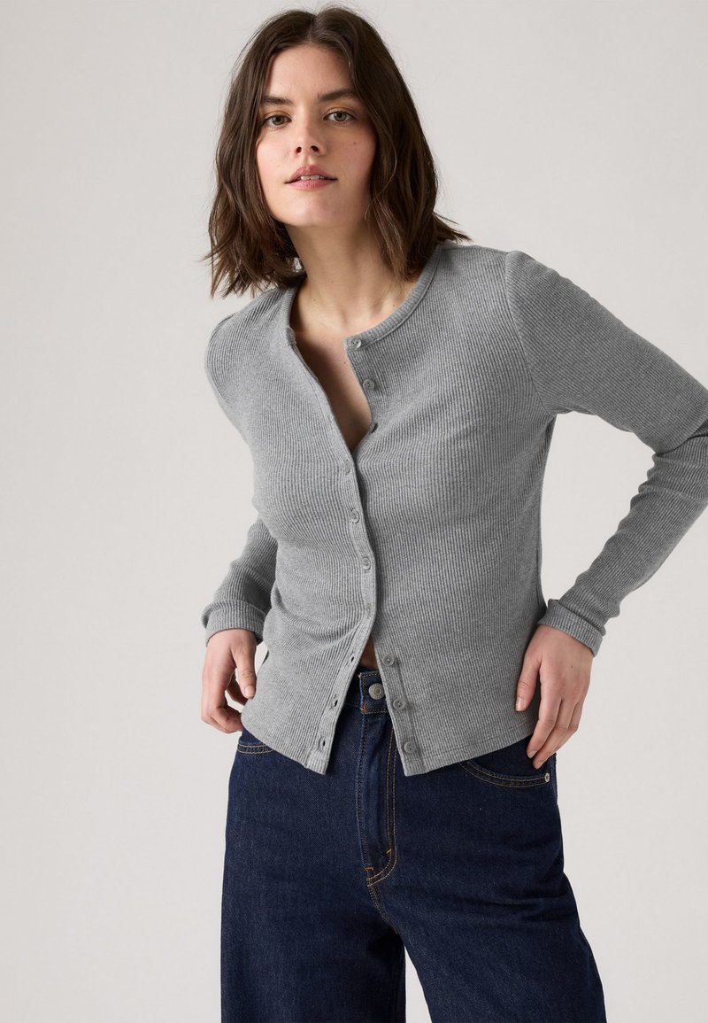 Grau gerippter Cardigan mit rundem Halsausschnitt, Knopfverschluss und langen Ärmeln, kombiniert mit blauen Jeans. Weiche Textur mit figurbetontem Schnitt.