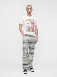 Homme portant un t-shirt blanc à motif graphique avec un aigle et du texte, un pantalon tie-dye gris-jaune et des baskets noires et blanches, debout sur un fond uni.