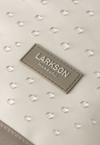 Béžová tkanina s vystúpenými vodnými kvapkami a obdĺžnikovou štítkou so stehovaniem, na ktorej je bielym písmom nápis „LARKSON HAMBURG“.