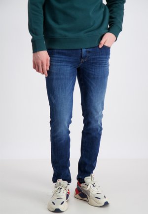 Jeans ajustados de un azul oscuro con un ligero desgastado, combinados con zapatillas en crema, rojo y azul marino, sobre un fondo liso.