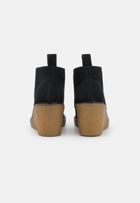 Botas de tobillo de ante negro con un diseño afilado, un tacón de cuña marrón texturizado y tiradores en la parte trasera para facilitar su uso.