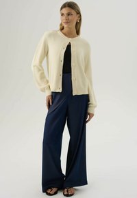 Donna che indossa un cardigan crema con bottoni sopra una maglia nera, pantaloni larghi blu navy e infradito nere, in piedi con una mano in tasca.