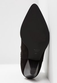 Zapato de ante negro de punta afilada con una suela de goma negra lisa. El texto en la suela dice "HECHO EN ESPAÑA" y la talla "37."