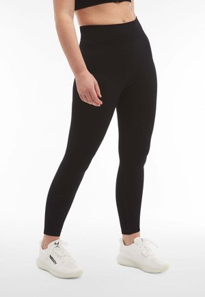 Leggings de sport noirs avec une taille haute, conçus en tissu lisse et extensible, associés à des chaussures de sport blanches.