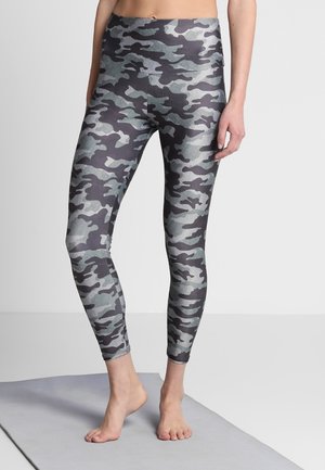Modelo con leggings ajustados de patrón de camuflaje, está descalza sobre una alfombrilla gris, destacando el diseño de las leggings contra un fondo neutral.