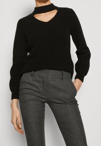 Kobieta ubrana w czarny sweter z długim rękawem i asymetrycznym wycięciem przy szyi oraz dopasowane spodnie w szarym jodełkowym wzorze, jedna ręka w kieszeni.