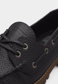 Chaussure bateau en cuir noir texturé avec détails à lacets et semelle en caoutchouc marron, vue en angle montrant les parties supérieure et latérale.