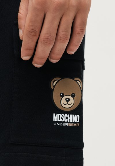 Χέρι που ακουμπάει σε μαύρο ύφασμα με γραφικό καφέ αρκούδας και κείμενο «MOSCHINO UNDERBEAR» κάτω από το πρόσωπο της αρκούδας.