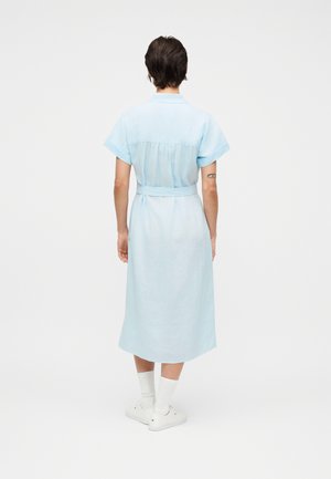 Rövid sötét hajú személy világoskék midi ruhában, fehér zokniban és fehér tornacipőben, hátával a néző felé áll egy fehér háttér előtt.