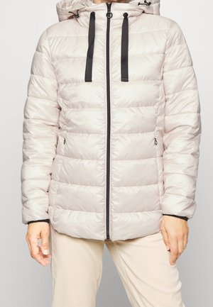 Lichtbeige gewatteerde donzen jas met zwarte ritssluiting en trekkoorden, gedragen met beige broek, handen ontspannen langs het lichaam.