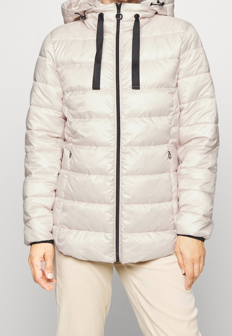 Veste matelassée beige clair avec fermeture éclair noire et cordons de serrage, portée avec un pantalon beige, les mains détendues le long du corps.