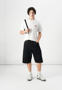Witte t-shirt met een kleine rode graphic, gecombineerd met knielange donkere denim shorts en witte sneakers met zwarte strepen. Model houdt een zwarte band vast.