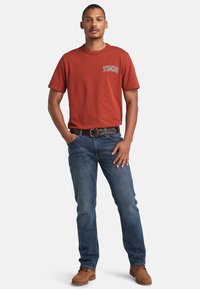 Rust-colored cotton t-shirt with "TIMBS" graphic, paired with blue denim jeans and brown suede boots, featuring a black belt.