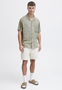 Lys grøn kortærmet skjorte med palmen mønster, parret med beige shorts. Model iført hvide sokker og brune slidesandaler.
