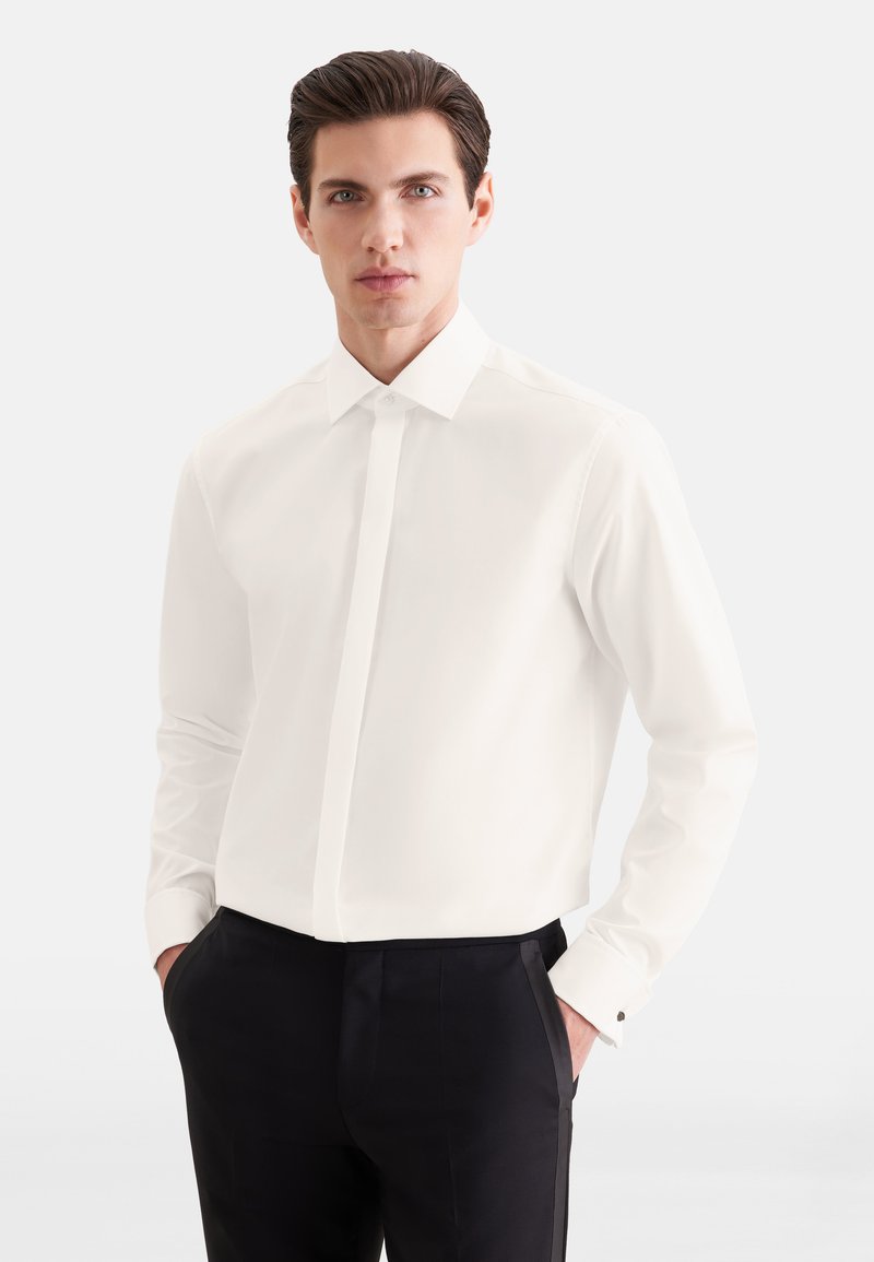 Homme portant une chemise blanche avec des boutons cachés et un pantalon habillé noir, debout les mains dans les poches sur fond blanc.