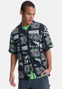 Μπλούζα με κοντά μανίκια σε μαύρο χρώμα με αφηρημένα λευκά και γκρι μοτίβα, διακοσμημένη με πράσινες γραφικές λεπτομέρειες και άνετη γραμμή. Από κάτω φοράει πράσινο T-shirt.