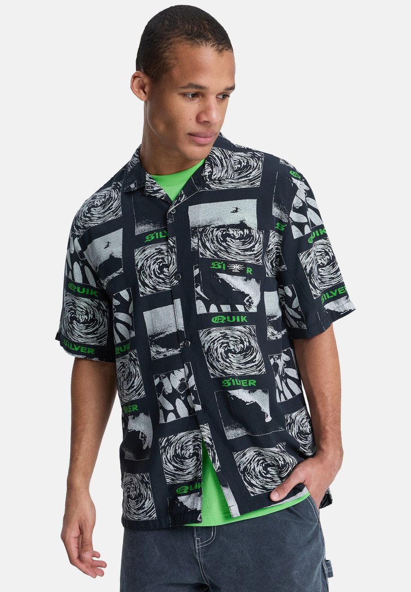 Μπλούζα με κοντά μανίκια σε μαύρο χρώμα με αφηρημένα λευκά και γκρι μοτίβα, διακοσμημένη με πράσινες γραφικές λεπτομέρειες και άνετη γραμμή. Από κάτω φοράει πράσινο T-shirt.