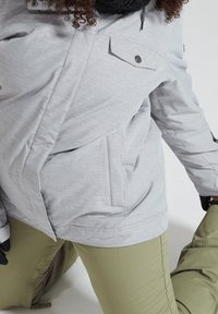 Personne portant une veste d'hiver gris clair, un pantalon vert olive, des gants noirs et une écharpe noire, à genoux sur un genou devant un fond blanc.