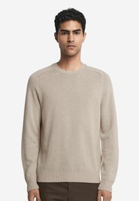 Beige gestrickter Pullover, Rundhalsausschnitt, gerippte Bündchen und Saum, glatte Textur, lange Ärmel, taillierte Silhouette. Getragen mit dunklen Hosen.