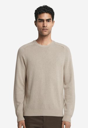 Pull en tricot beige, col rond, poignets et ourlet côtelés, texture lisse, manches longues, silhouette ajustée. Porté avec un pantalon sombre.