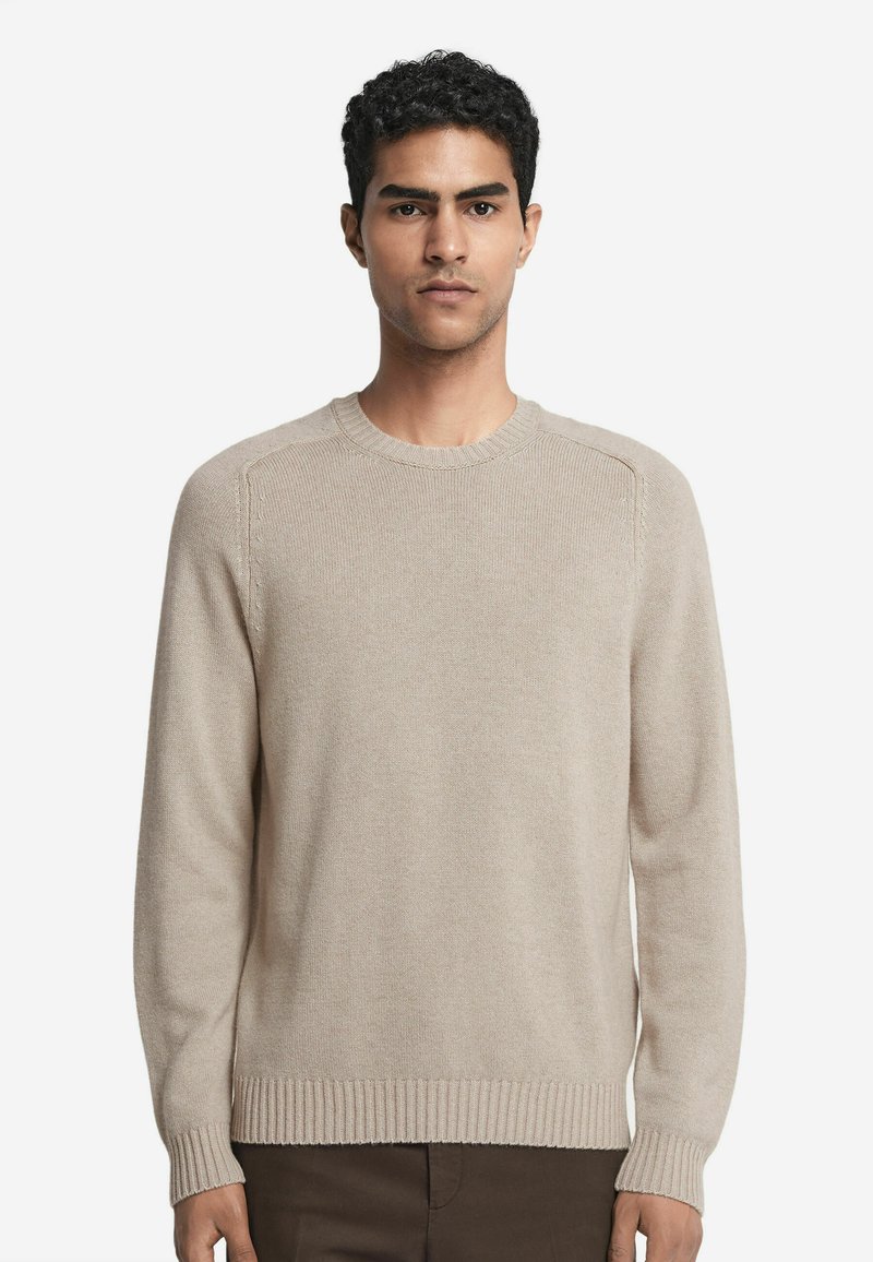 Beige gestrickter Pullover, Rundhalsausschnitt, gerippte Bündchen und Saum, glatte Textur, lange Ärmel, taillierte Silhouette. Getragen mit dunklen Hosen.