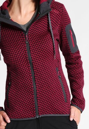 Kvinde iført rød og sort hundetandsmønstret jakke med lynlås, hætte og lynlåslommer foran og på ærmet.