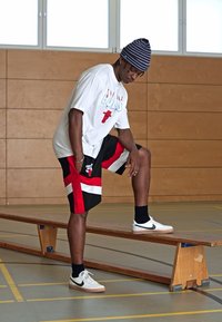 Bela majica s rdečim grafičnim motivom Chicago Bulls, črnordeče kratke hlače s črtami in bele superge s črnimi poudarki, sedi na klopi.