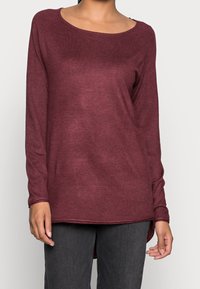 Weinrotes Langarmshirt mit weitem Ausschnitt, gerippter Textur und einem leicht verlängerten Rückensaum, kombiniert mit dunkelgrauen Jeans.