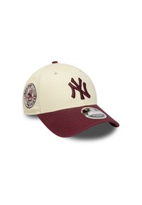 Baseball cap with a cream body, maroon visor, and embroidered NY logo. Includes a round emblem on the side and a label on the brim.