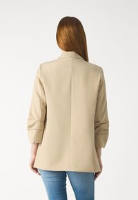 Beige taillierter Blazer mit Drei-Viertel-Ärmeln, glatter Textur und einer einzigen Rückennaht, kombiniert mit blauen Jeans.
