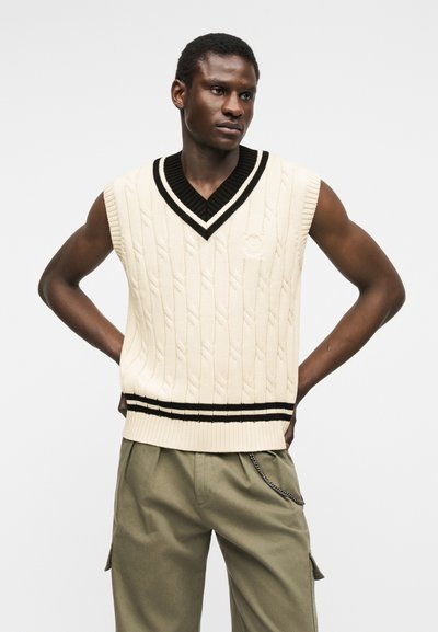 Νεαρός άνδρας που φοράει αμάνικο πουλόβερ μπεζ πλεκτό με μαύρο περίγραμμα και χακί παντελόνι cargo, στέκεται με τα χέρια στη μέση.