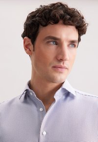 Camicia bianca con bottoni, decorata con un motivo geometrico blu navy, colletto strutturato e una consistenza in cotone morbido. Dettagli degni di nota includono bottoni a contrasto.
