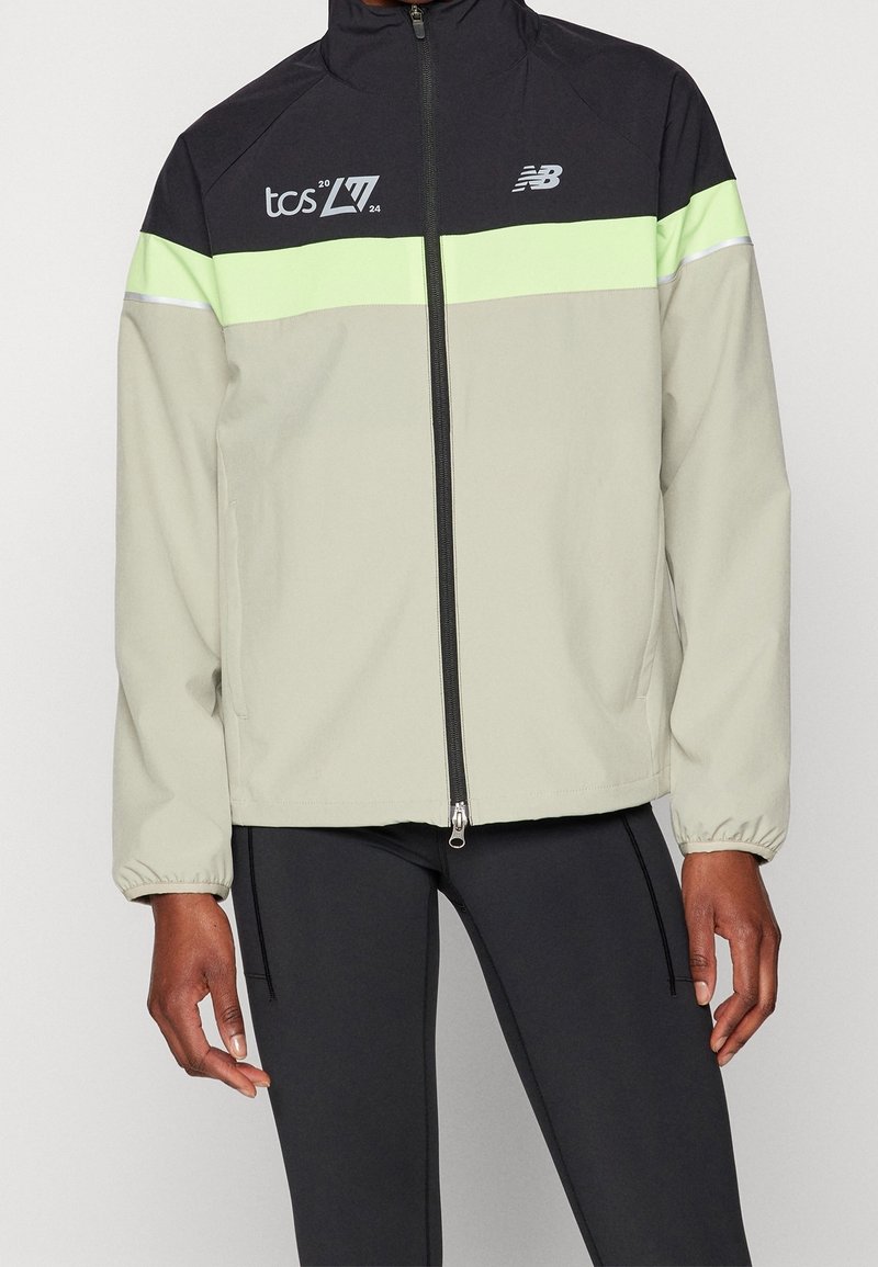 Veste zippée noire et beige avec bande vert néon, logos "tcs 2024" et New Balance sur la poitrine, portée avec un pantalon noir.