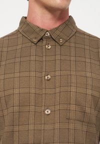 Homme portant une chemise à carreaux marron boutonnée avec un col boutonné et une poche poitrine visible.