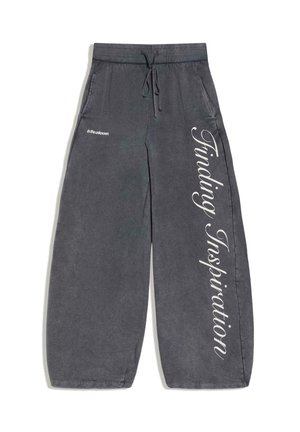 Sötétszürke, széles szárú melegítőnadrág húzózsinóros derékkal, oldalzsebekkel, valamint fehér szöveggel, amelyen "in the unknown" és "Finding Inspiration" feliratok találhatók a lábszárakon.