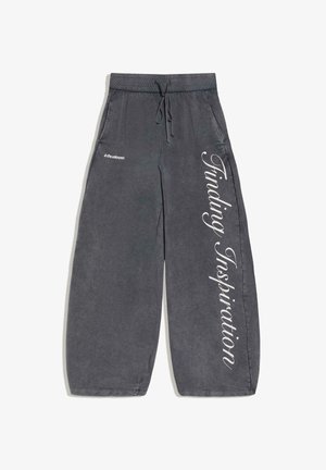 Donkergrijze sweatpants met wijde pijpen, een trekkoord in de taille, zijzakken en witte tekst "in the unknown" en "Finding Inspiration" op de pijpen gedrukt.