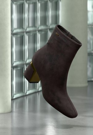 Mørkebrun ruskind ankle-støvle med blokhæl og gulddetalje, vist foran en glasblokvæg og poleret gulv.
