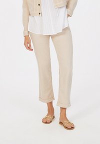 Beige Leinenhose mit aufgerolltem Saum, kombiniert mit einer blauen gestreiften Bluse. Dazu flache Sandalen mit goldenen Riemen und brauner Sohle.