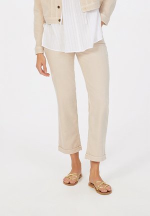 Beige Leinenhose mit aufgerolltem Saum, kombiniert mit einer blauen gestreiften Bluse. Dazu flache Sandalen mit goldenen Riemen und brauner Sohle.