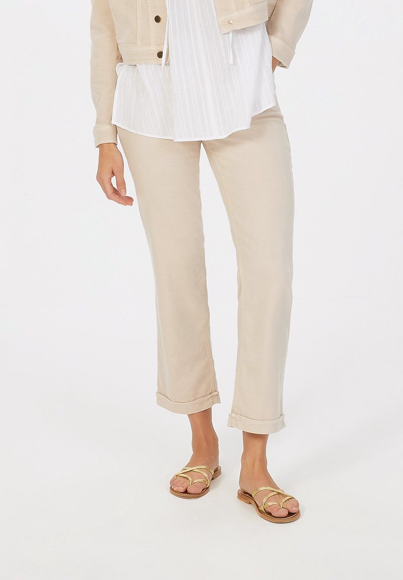 Beige Leinenhose mit aufgerolltem Saum, kombiniert mit einer blauen gestreiften Bluse. Dazu flache Sandalen mit goldenen Riemen und brauner Sohle.