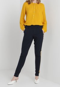 Chemisier jaune à col rond, associé à un pantalon noir cintré avec des rayures dorées sur les côtés. Le modèle porte des chaussures à talons blanches.
