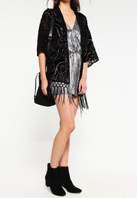 Kimono de encaje negro con patrones intrincados, llevado sobre un vestido plateado de flecos. Botines negros y un pequeño bolso negro completan el look.