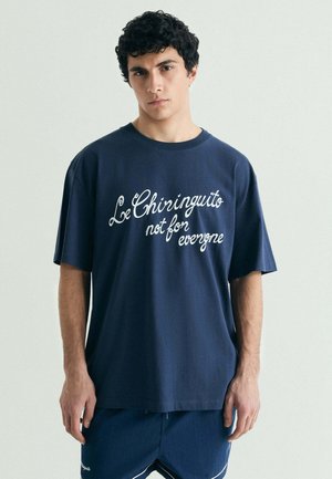 Joven con cabello rizado que lleva una camiseta azul marino con texto blanco "Le Chiringuito not for everyone" y pantalones cortos azules marinos a juego.