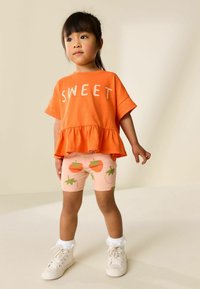 Narancssárga rövid ujjú felső "SWEET" felirattal, peplum szegéllyel, rózsaszín rövidnadrággal, amely paradicsom mintákat visel, és fehér csipkével díszített zoknikkal párosítva.