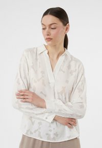 Witte blouse met een subtiel gemêleerd patroon, kraagloos ontwerp en lange mouwen, met knoopmanchetten. Lichtgewicht en semi-transparante stof.